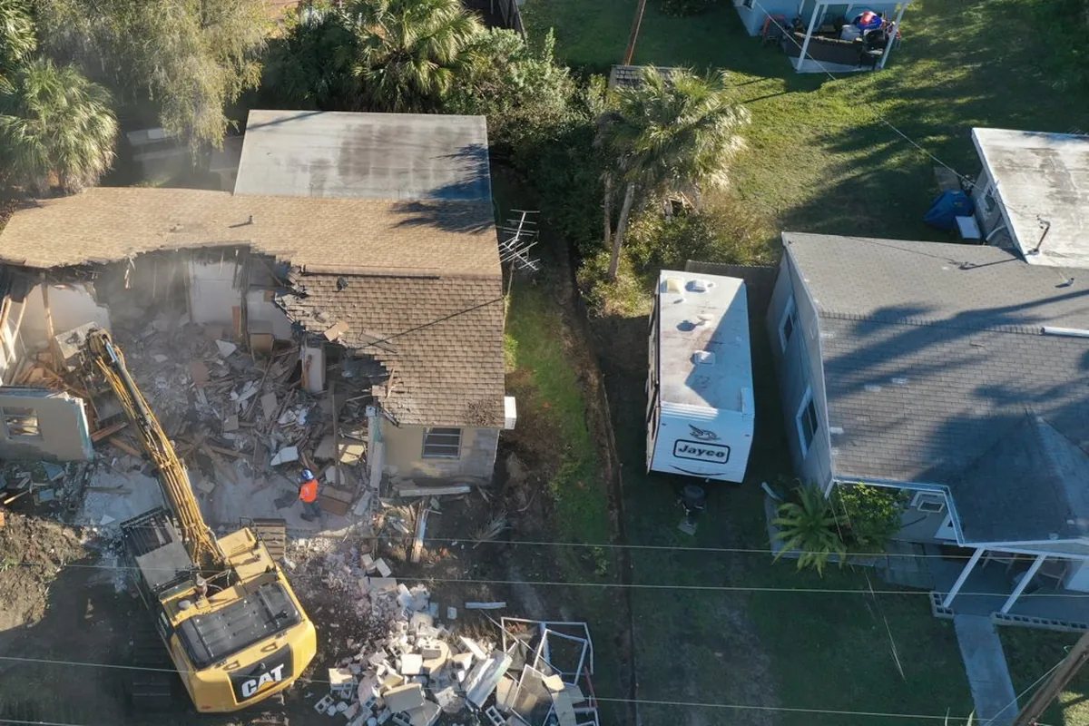 Excavator tearing down a small house on a residential lot, with block rubble piled beside it