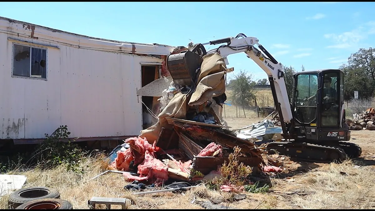 Mini excavator tearing the end off an old single wide mobile home, with insulation and debris piled in front