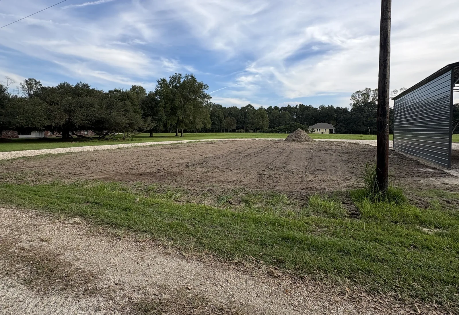Freshly graded dirt pad with a spoil pile and a gravel drive beside a metal building