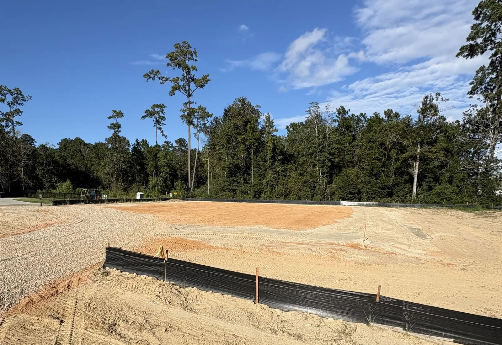 Compacted sand pad with track marks behind a silt fence, pines and houses behind it